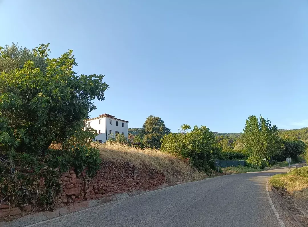 Una carretera rural pavimentada curva hacia la derecha, con un edificio blanco de varias plantas en una ladera seca y herbosa a la izquierda, rodeado de árboles bajo un cielo azul claro.